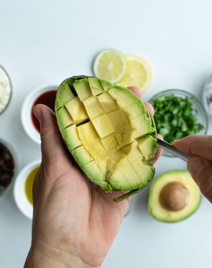 hand holding avocado and scooping cubes of avocado out of the skin with ingredients for greek guacamole in the background