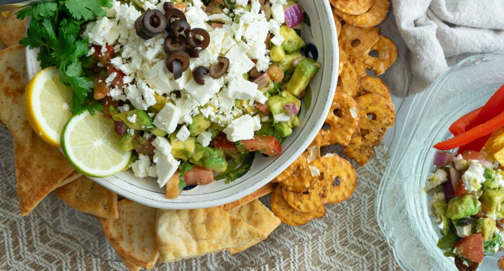 greek guacamole in a white bowl garnished with cilantro and lime topped with feta cheese and black olives, platter has pretzels, pita chips, and tortilla chips