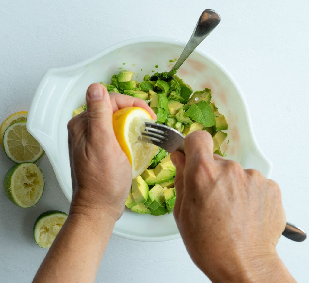 squeezing lemon over diced avocado in a white bowl with a silver spoon in the bowl on a gray countertop