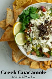 greek guacamole in white bowl surrounded by pita chips, pretzel crisps, and tortilla chips