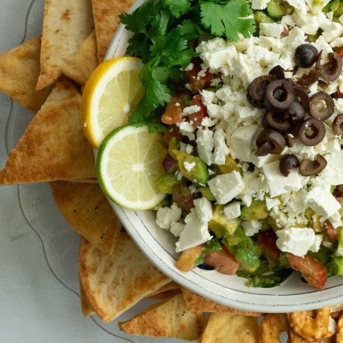 greek guacamole in white bowl surrounded by pita chips, pretzel crisps, and tortilla chips