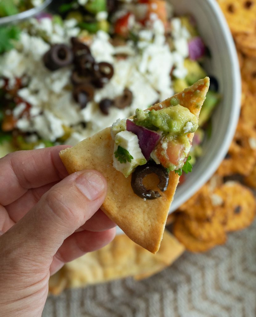 hand holding a pita chip with greek guacamole on it with bowl of greek guacamole in the background with platter of dippers 