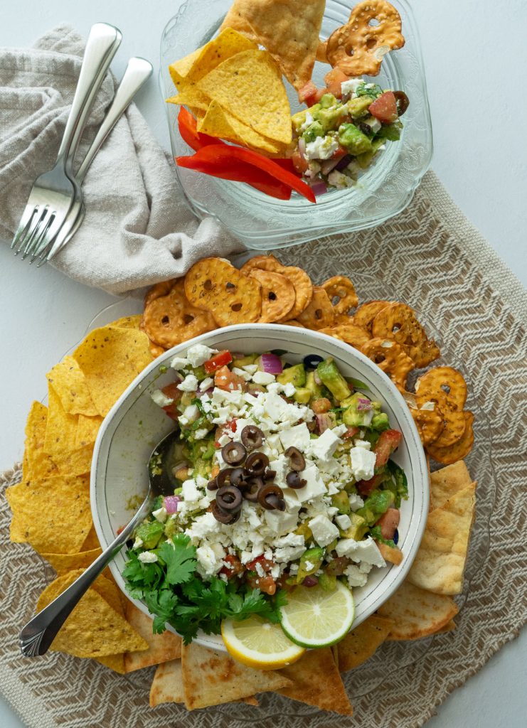 display of greek guacamole in a serving dish surrounded by pretzels, pita chips, and tortilla chips with  plates, forks and napkins above the guacamole platter