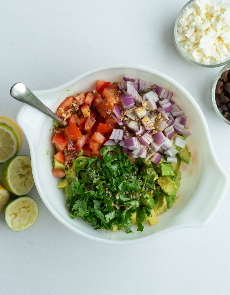 diced tomatoes, diced red onion, chopped cilantro over diced avocados  in a medium white mixing bowl with a silver spoon sticking out of the left side.  squeezed limes and lemons off to the left of the bowl, feta cheese and black olives in the upper righthand corner. 
