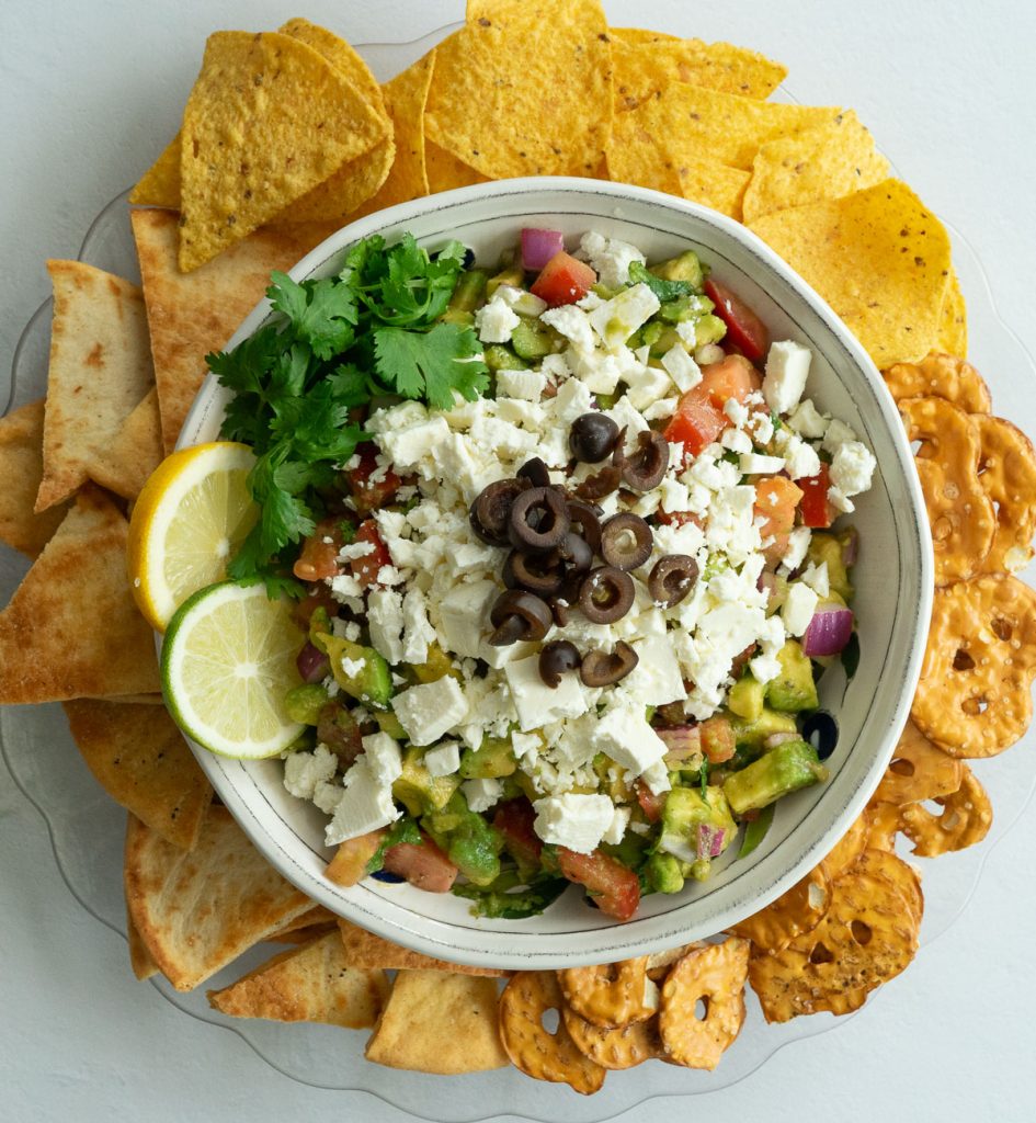 greek guacamole in white dish with feta cheese and black olives on the top garnished with lime and cilantro and a platter of pita chips, pretzels, and tortilla chips