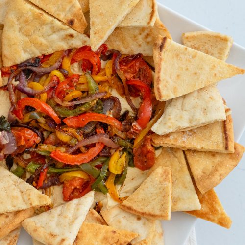 roasted vegetables and hummus dip surrounded by pita chops on a white platter gray background
