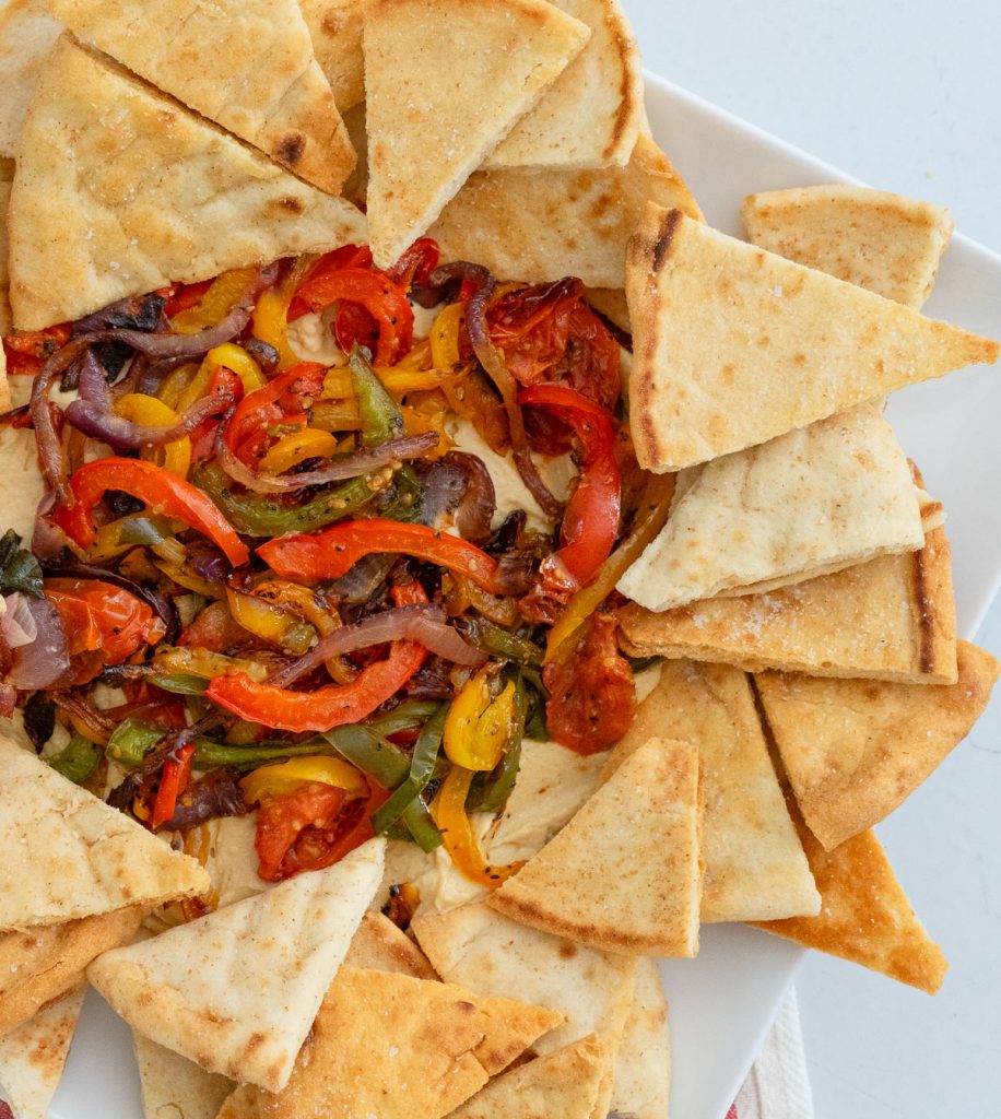 roasted vegetables and hummus dip surrounded by pita chops on a white platter gray background