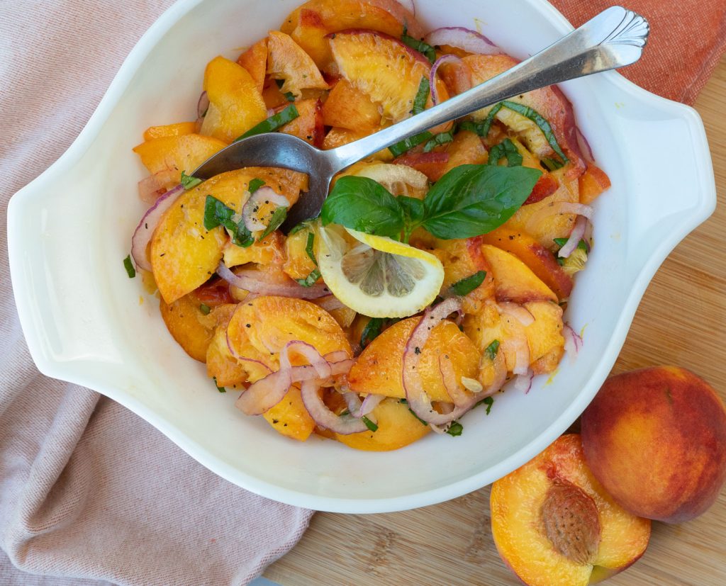 white bowl with peach basil salad in it, silver serving spoon, garnished with basil and lemon with a whole peach sliced in half in the bottom right corner of the photo