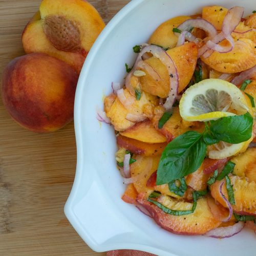 white bowl with peach basil salad in it garnished with lemon slice and basil leaves on a wood countertop a peach sliced in half on countertop in upper left hand corner of picture
