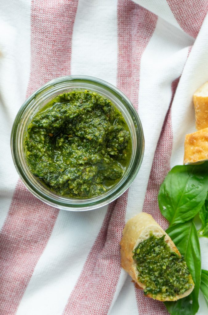 roasted garlic pesto in a glass mason jar with a piece of bread with pesto spread on it red and white kitchen towel in the background