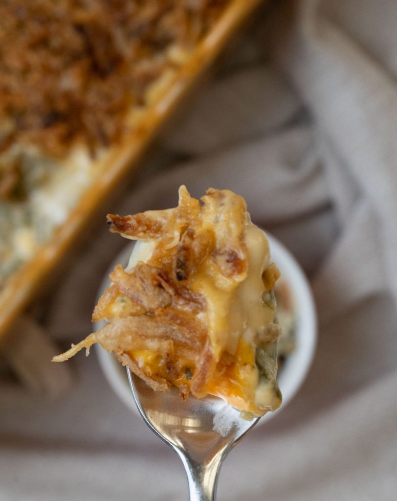 perfect spoon full of green bean casserole with dish of casserole in the back ground on a tan background