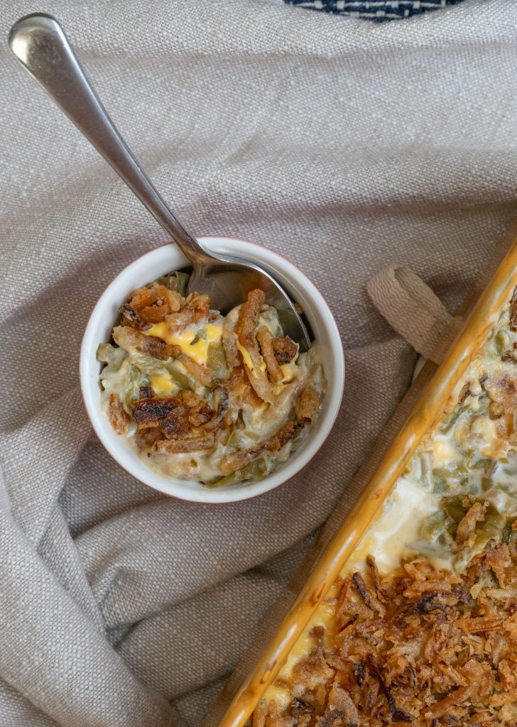 small bowl of green bean casserole sitting next to dish of casserole on a tan napkin with a silver spoon in small bowl 