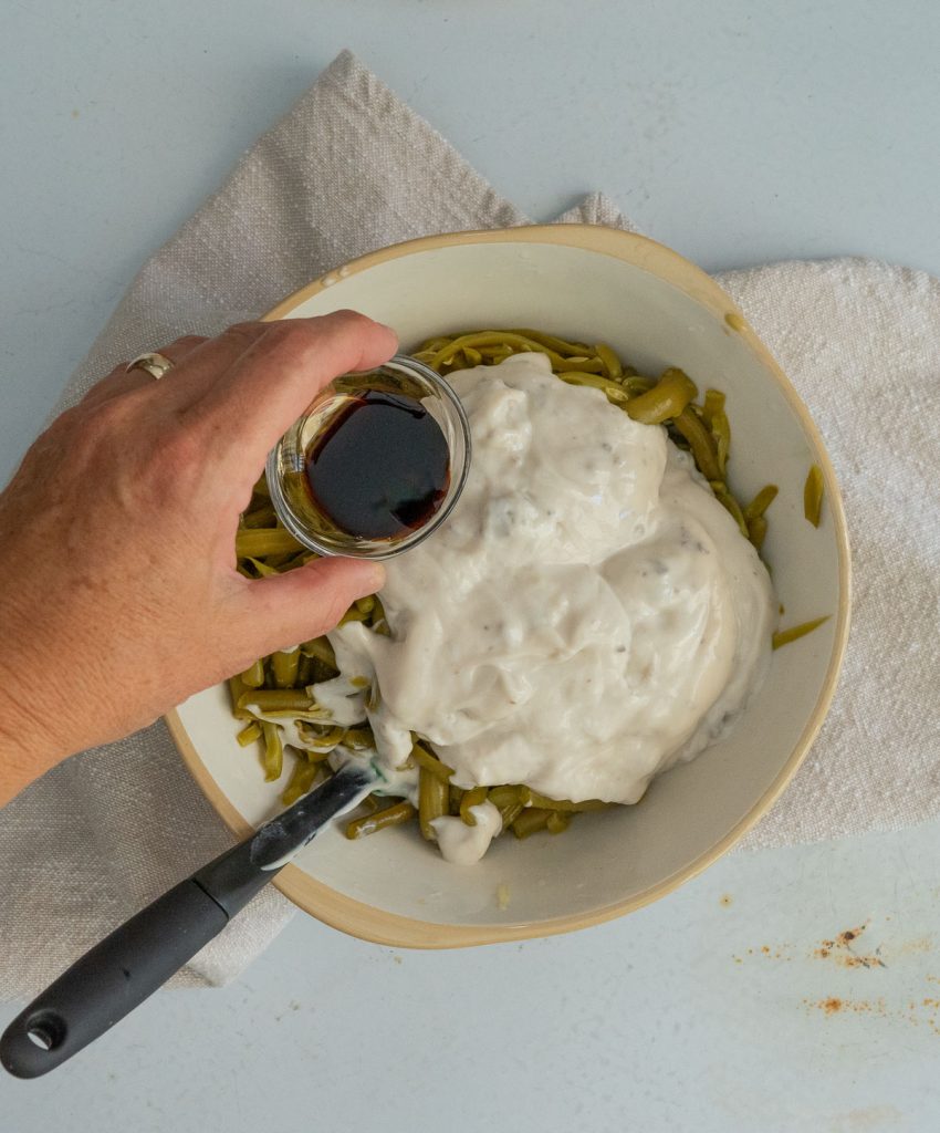 bowl with green bean, cream of mushroom soup in it, with a spatula at 7 o'clock and a hand pouring in the soy sauce