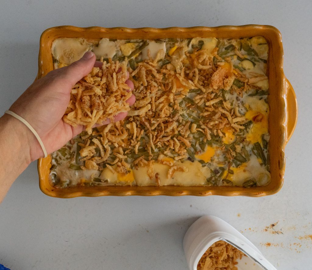 hand coming from the left sprinkling french fried onions over the casserole