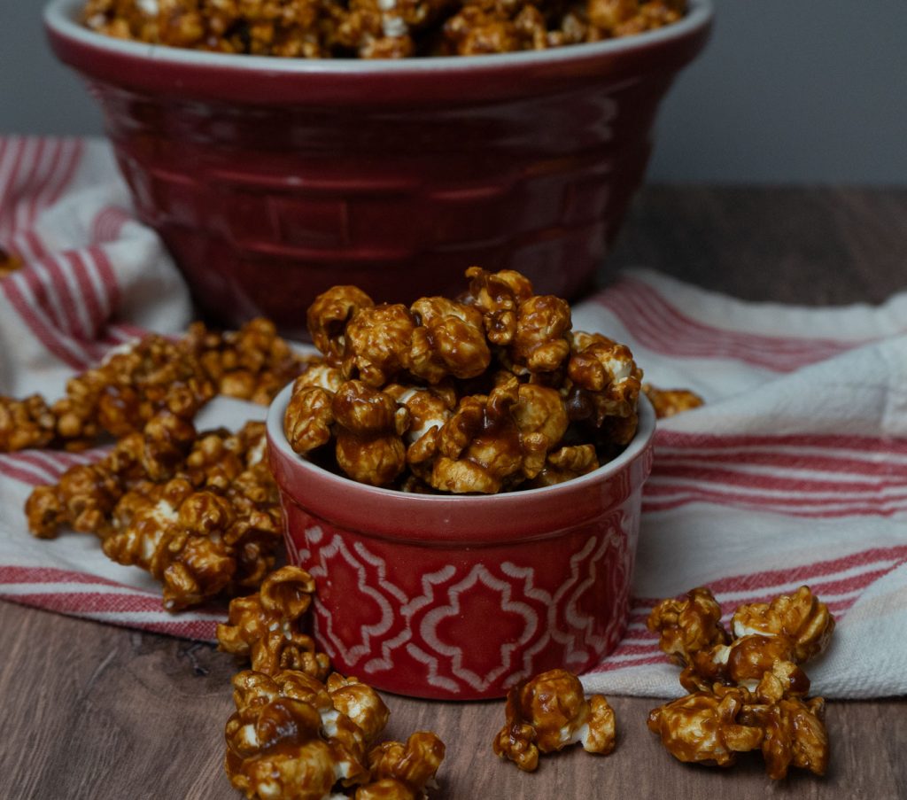 small red dish filled with gingerbread caramel corn with caramel corn all around the dish, larger bowl of gingerbread caramel corn in background