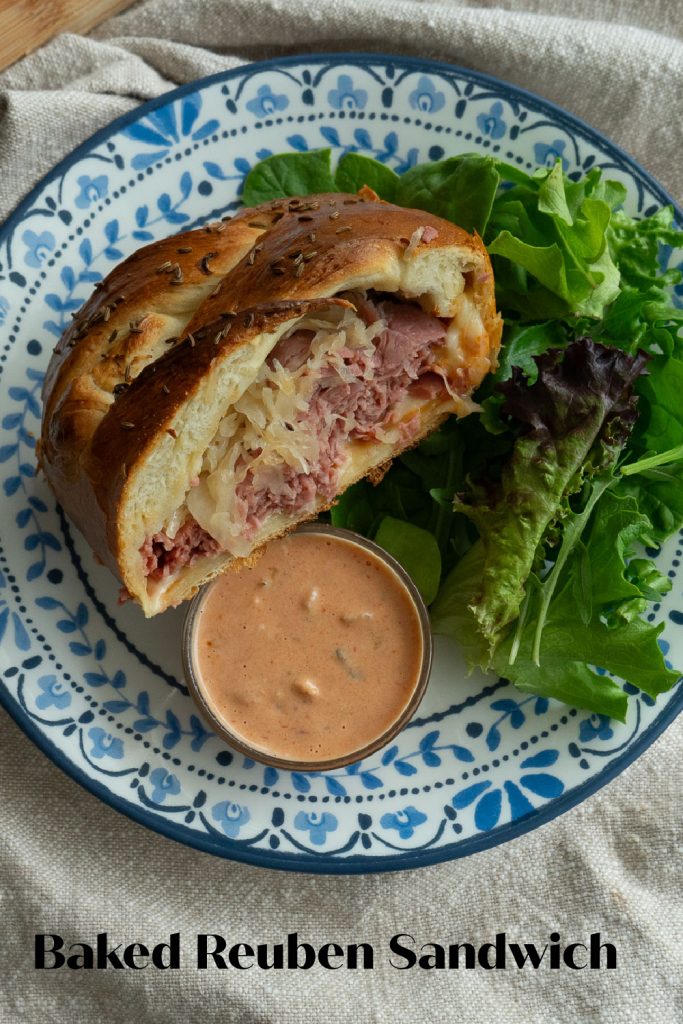 slice of baked reuben sandwich on a plate with blue edges with a side of thousand island dressing in a small glass dish and a portion of greens on the side