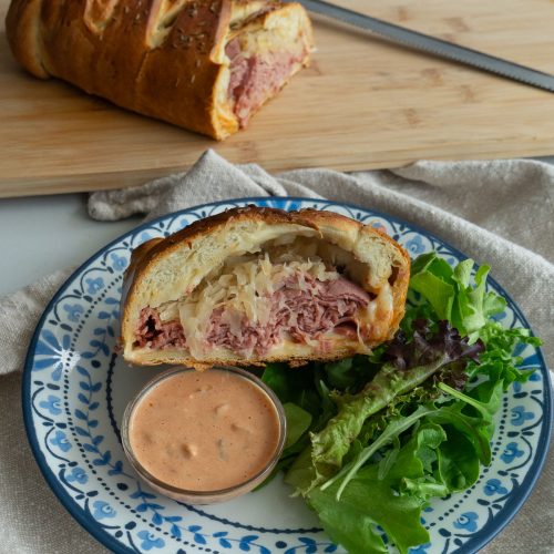 baked reuben sandwich on wood cutting board, slice of the sandwich is on a plate with a small dish of thousand island dressing and a salad.