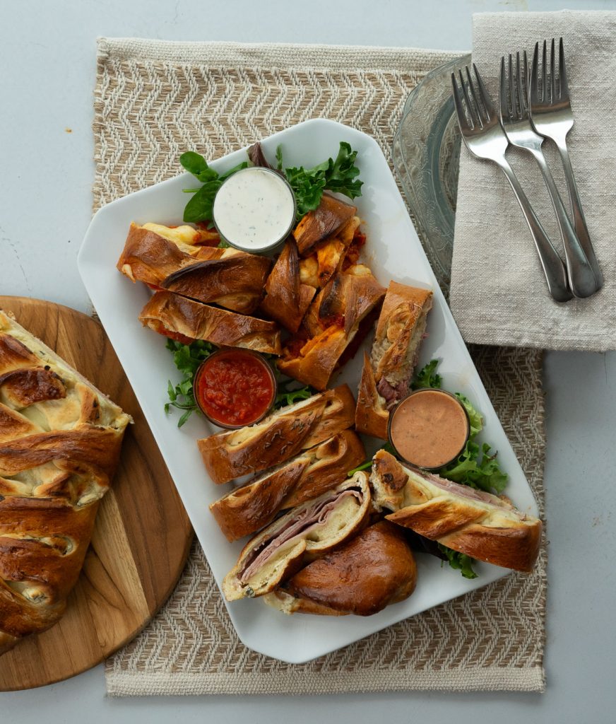 White Platter of baked sandwiches with small dishes of dipping sauces forks and napkins are placed in the upper right hand corner in the lower lefthand corner is a wood cutting board with an uncut baked sandwich on it