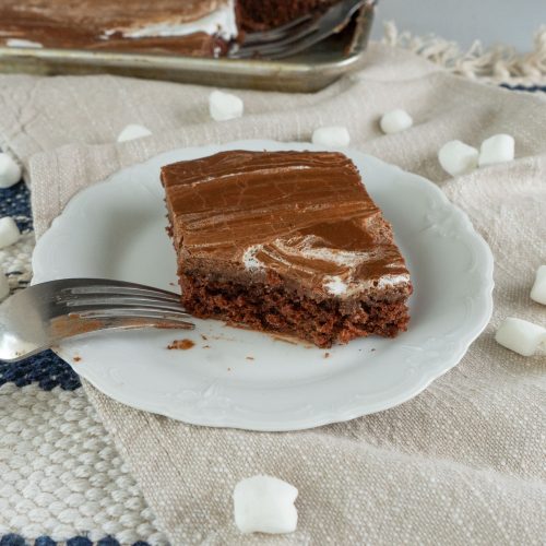 one square piece of michigan style texas sheet cake on a white plate with sheet cake in the background and a fork resting on the plate, small marshmallows are strewn around on a beige nakin, place pat