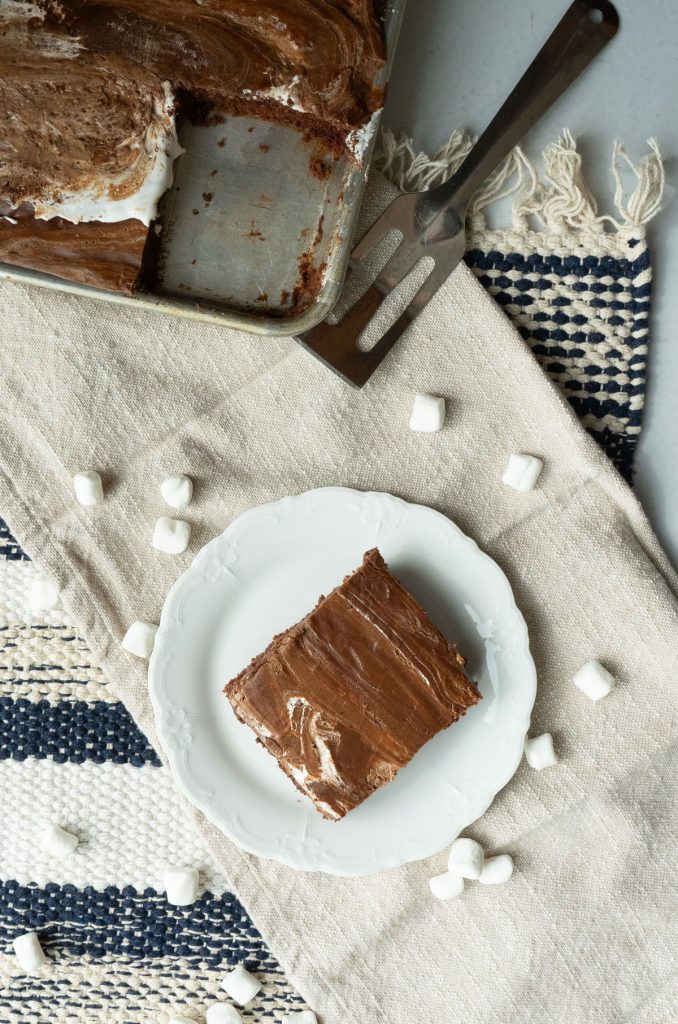 square of chocolate texas sheet cake on a white plate with cake pan in upper left hand corner, marshmallows strewn around