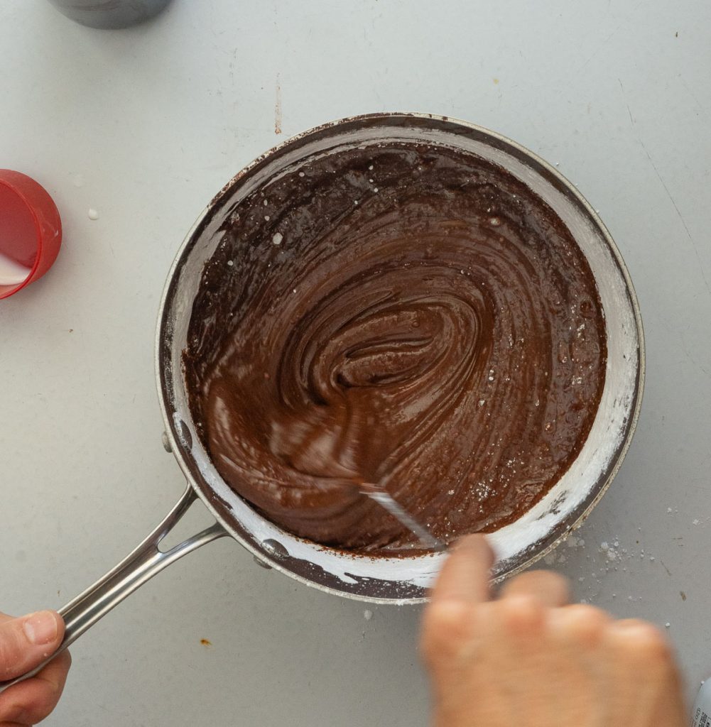 whisking fudge frosting in medium sized stainless steel sauce pan on a gray counter top