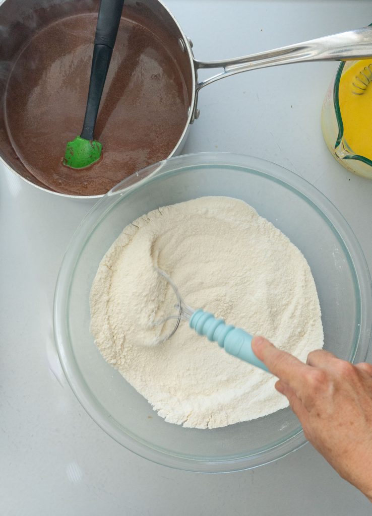 whisking together dry ingredients for texas sheet cake upper left hand corner has chocolate in sauce pan upper right hand corner has wet ingredients of egg and buttermilk