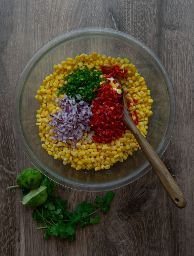 corn, red bell pepper, red onion, and jalepeno in a large glass bowl with a juiced lime and cilantro leaves in the lower left hand counter