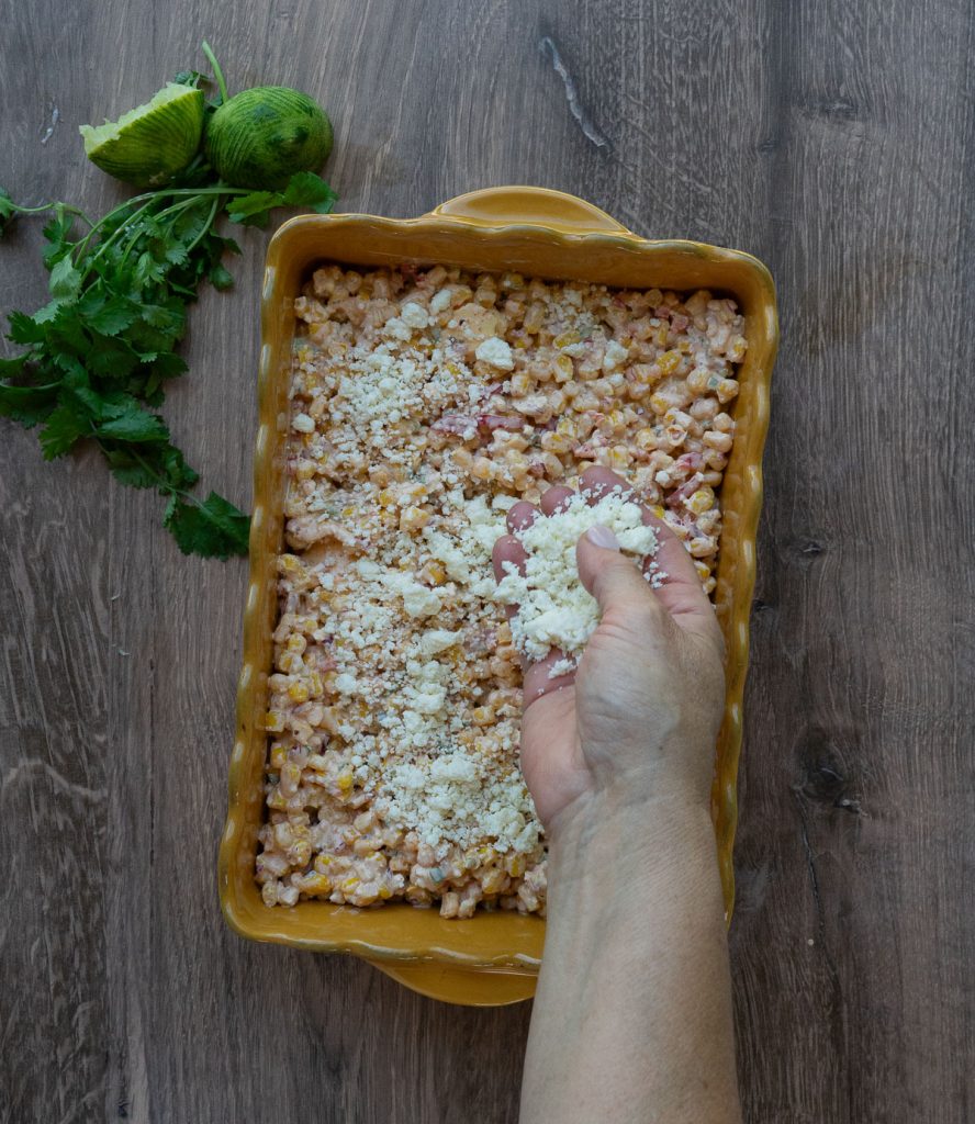 mexican corn casserole in a rectangle yellow baking dish on a wood countertop with cilantro and a lime in the upper left hand corner of the photo. A hand is sprinkling cheese over the top of the casserole