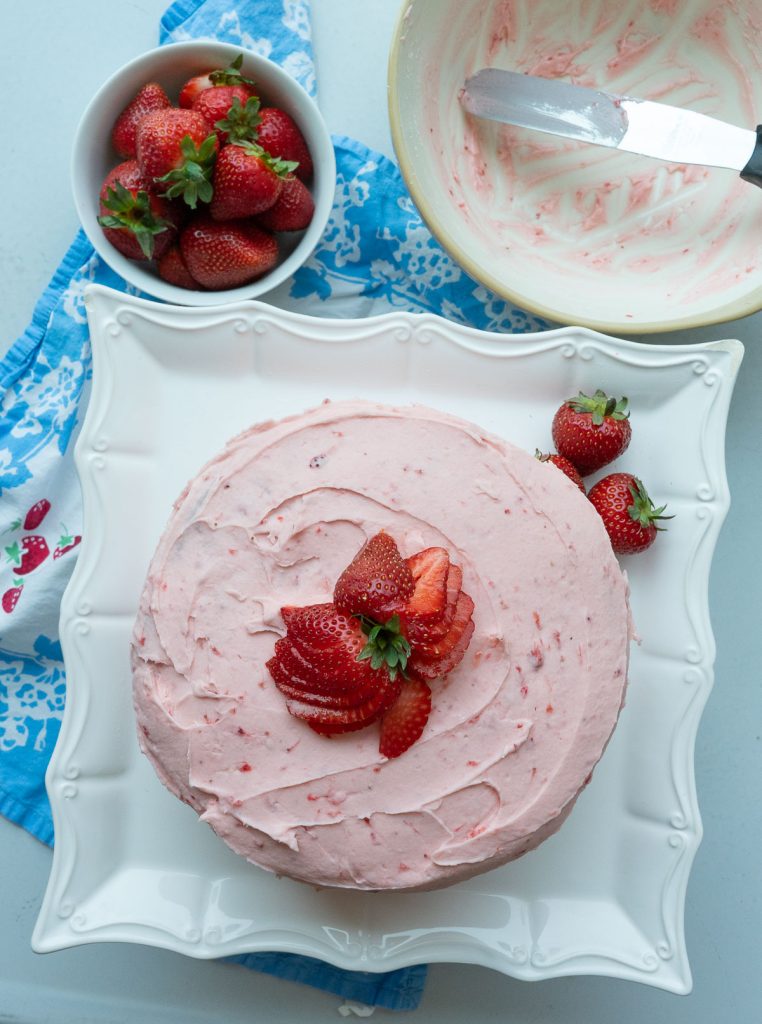 top view of strawberry cake with a bowl of fresh whole strawberries in the upper left hand corner, and an empty frosting bowl in upper rt hand corner