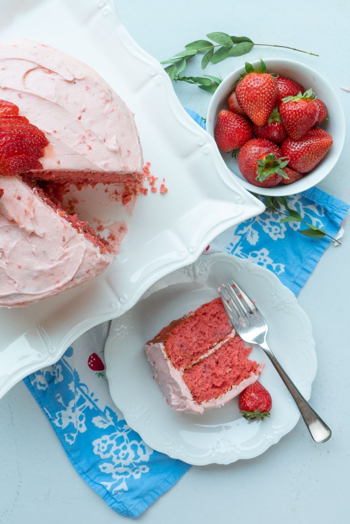 strawberry cake with a slice cut out. white plate with a piece of strawberry cake  on it with a fork to the side, bowl of whole strawberries in the upper right corner