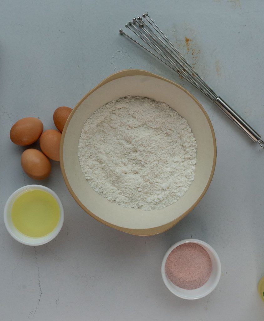 cake mix in a large mixing bowl, small bowl of vegetable oil off to the left, eggs, and a small white bowl with strawberry jello in it, whisk laying on table to the upper right 