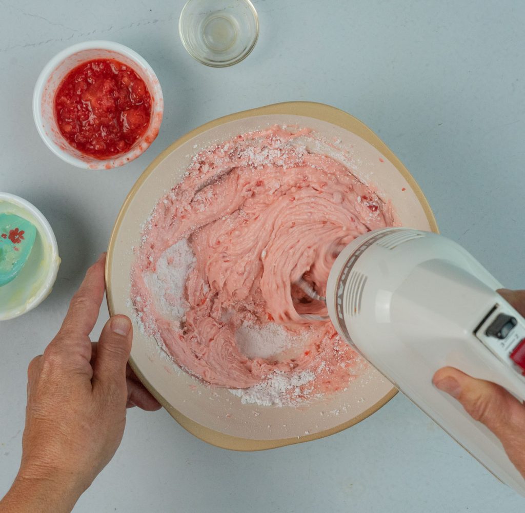 making strawberry frosting with a hand mixer , bowl of smashed strawberries in upper left hand corner 