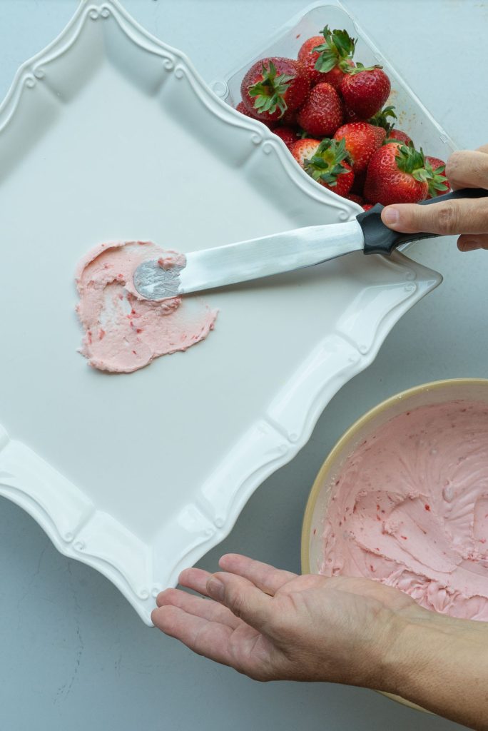 frosting the strawberry cake on a white pedistal stand fresh strawberries in the upper right hand corner and a bowl of frosting in lower right hand corner