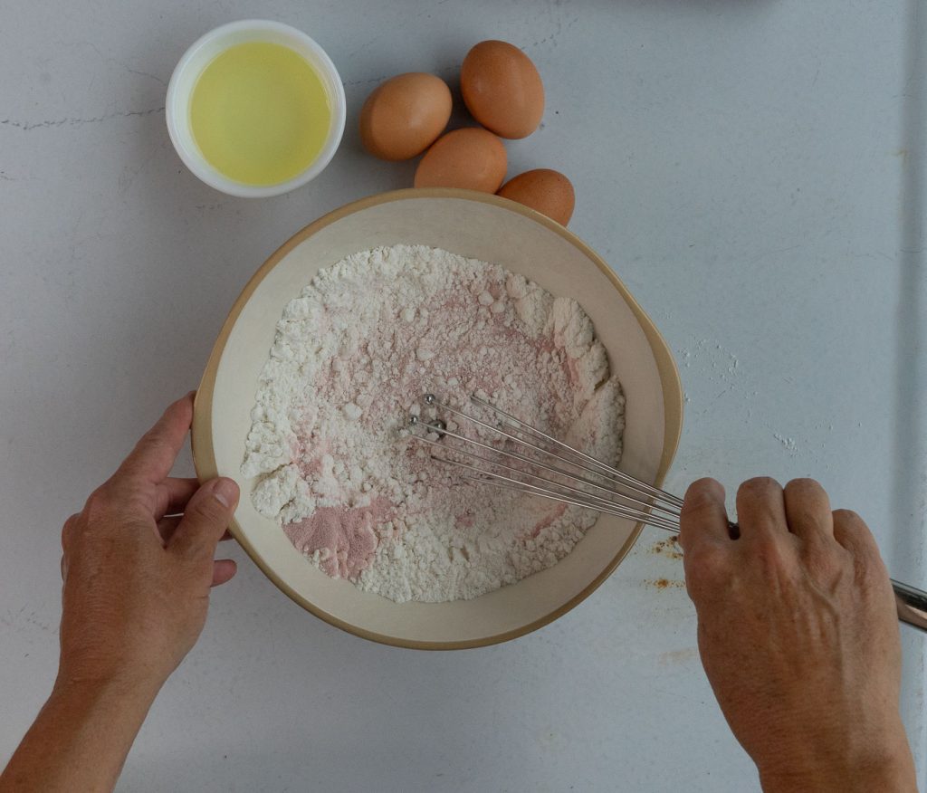 mixing strawberry jello into the white cake mix in large bowl.  eggs and vegetable oil is in the upper left hand corner of the photo