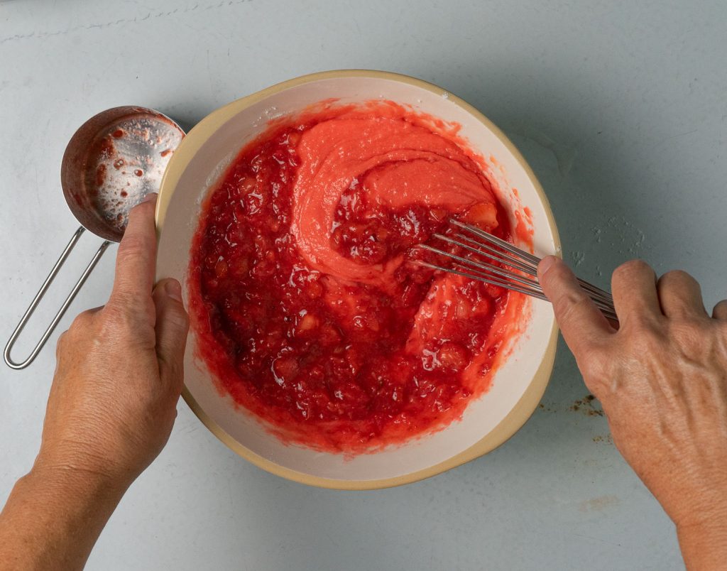 Adding fresh strawberries to the cake batter, hands are holding bowl from the left and whisking on the right