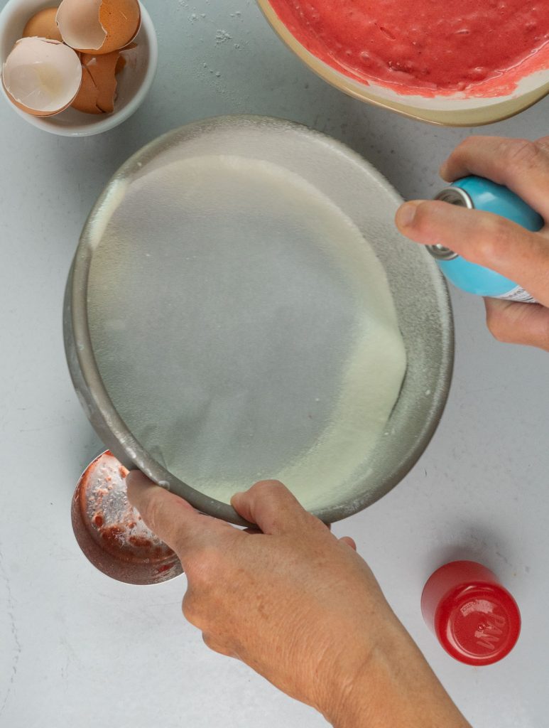 preparing round cake pans for baking strawberry cake. hand holding round cake pan, while spring with non stick baking spray with other hand