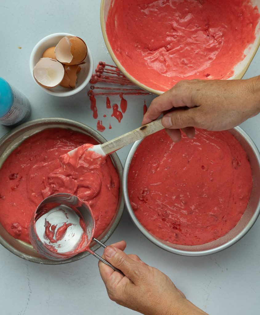 filling round cake pans with strawberry cake batter. bowl of egg shells is in upper left hand corner 