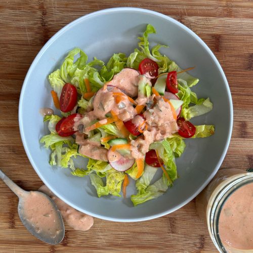 plate of salad with thousand island dressing on it, plate is on a wood cutting board with a bowl of dressing off to the side