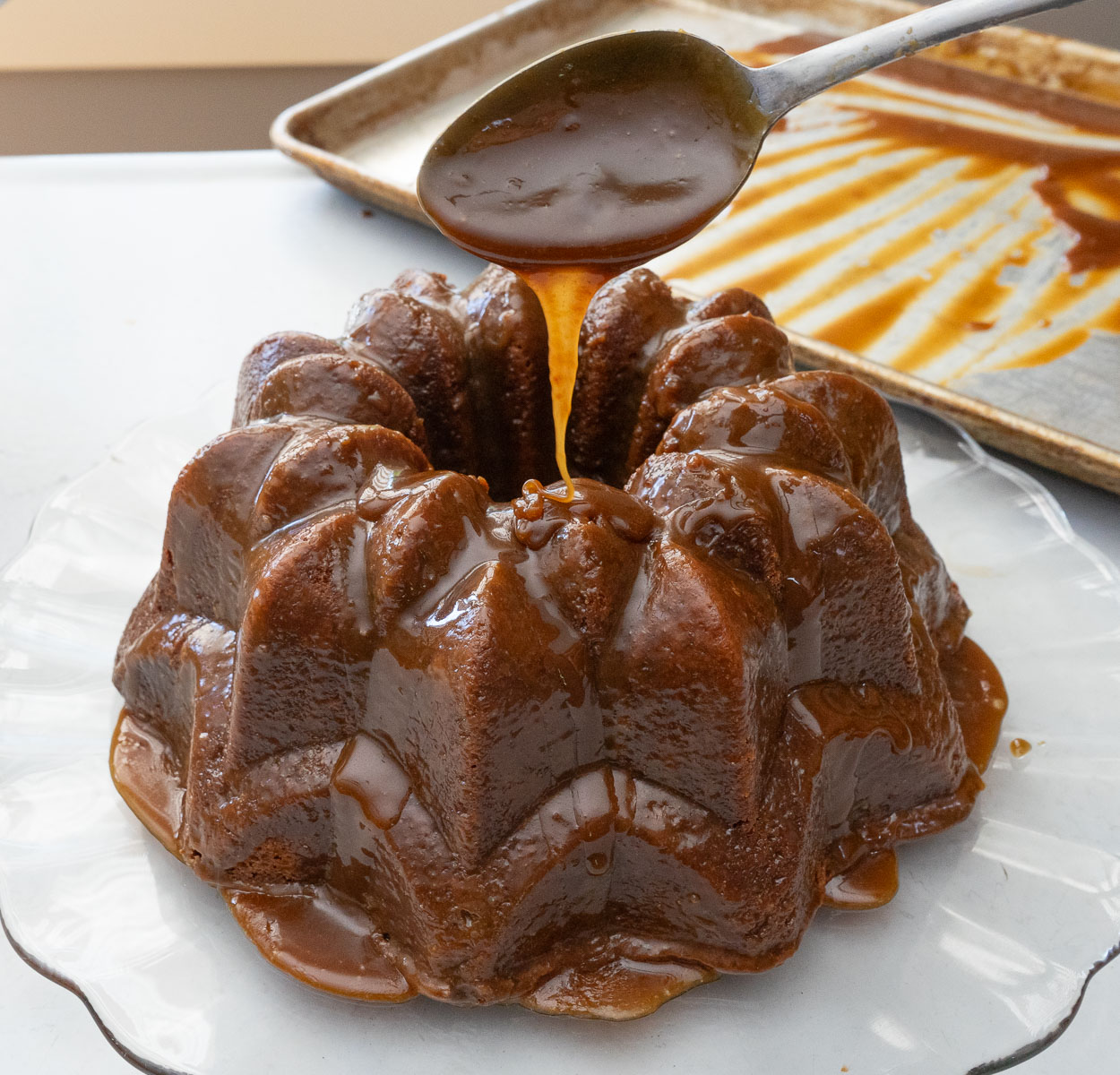 caramel toffee bundt cake on a glass cake plate with a spoon drizzling caramel glaze over the cake