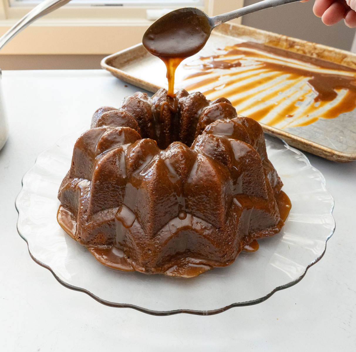 caramel toffee bundt cake on a glass cake plate with a spoon drizzling caramel glaze over top of the cake