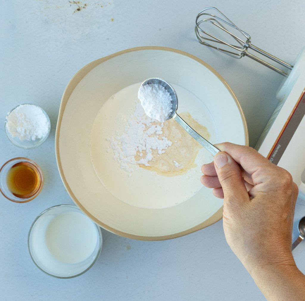 add vanilla and powdered sugar to heavy whipping cream hand mixer is off the right, glass dishes of heavy whipping cream, powdered sugar and vanilla on the left of bowl