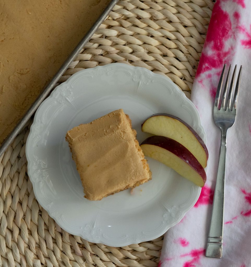 piece of cake on a white plate with slices of apples on a rattan placemat with fork and pink tie dyed napkin to the right of the plate 