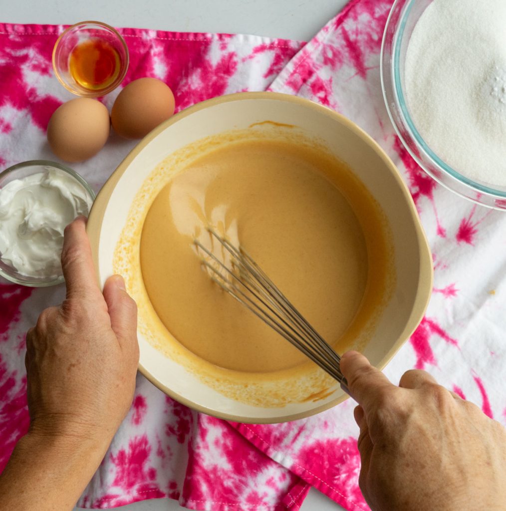 smooth mixture of peanut butter, butter, and water, being mixed with a whisk. bowl on a pink tie dyed cloth, with eggs, sour cream and dry ingredients surrounding the bowl