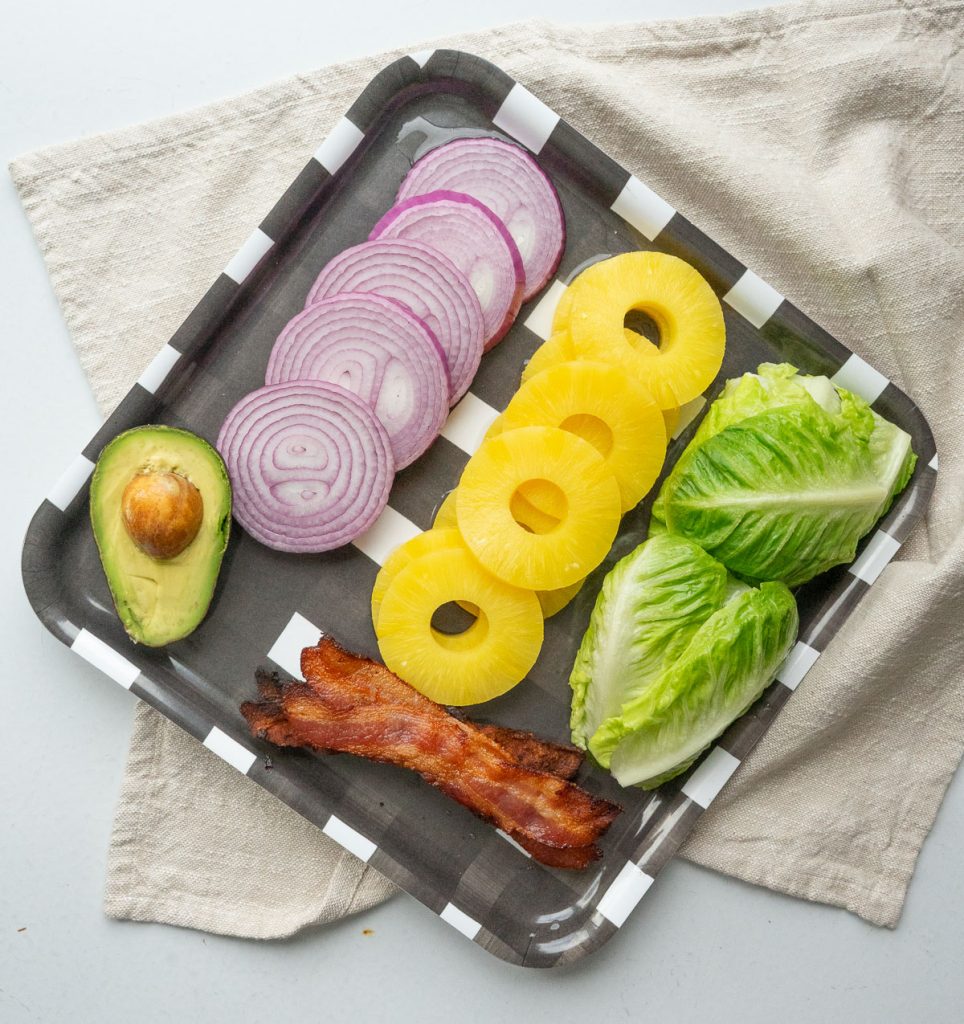 photo of avocado, red onion, pineapple, lettuce and bacon slices on a black and white checker board platter, on a tan napkin with gray background