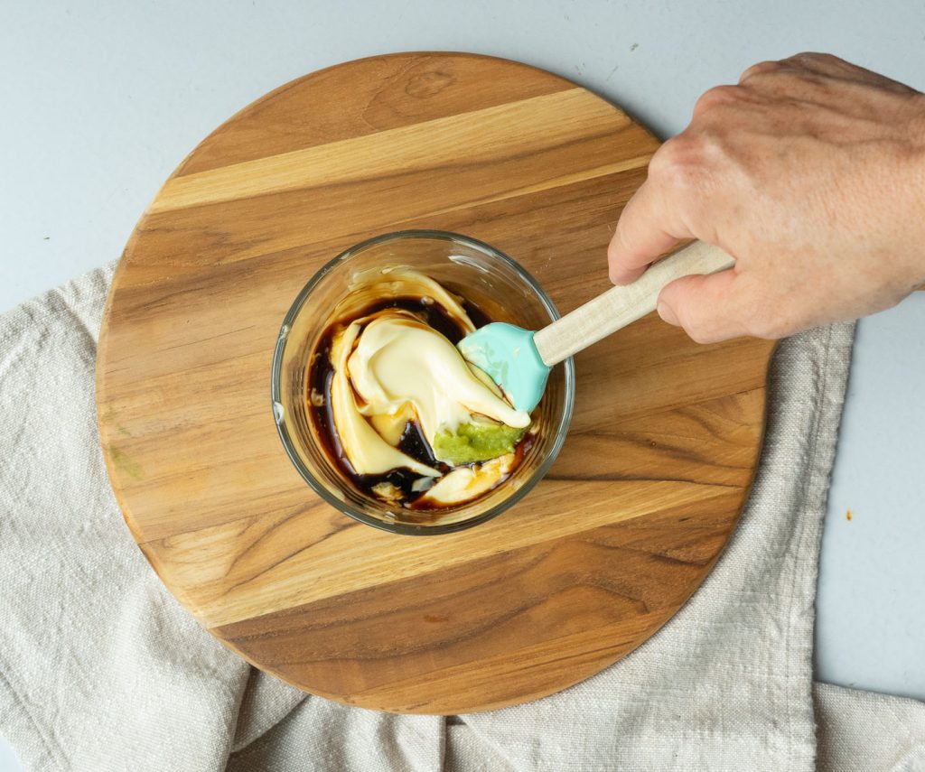 combining sauce ingredients together in a glass bowl with small spatula on a round wood cutting board