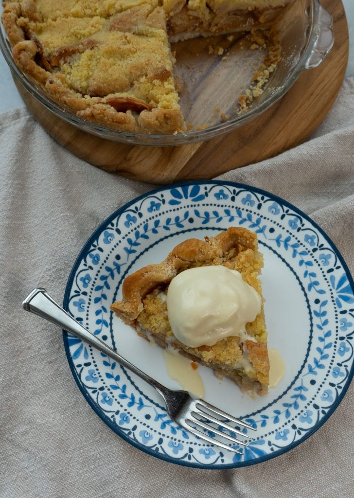 slice of dutch apple pie with ice cream on top. pie is on a white plate rimmed in blue with a fork on the plate. Whole pie is sitting above the piece of pie