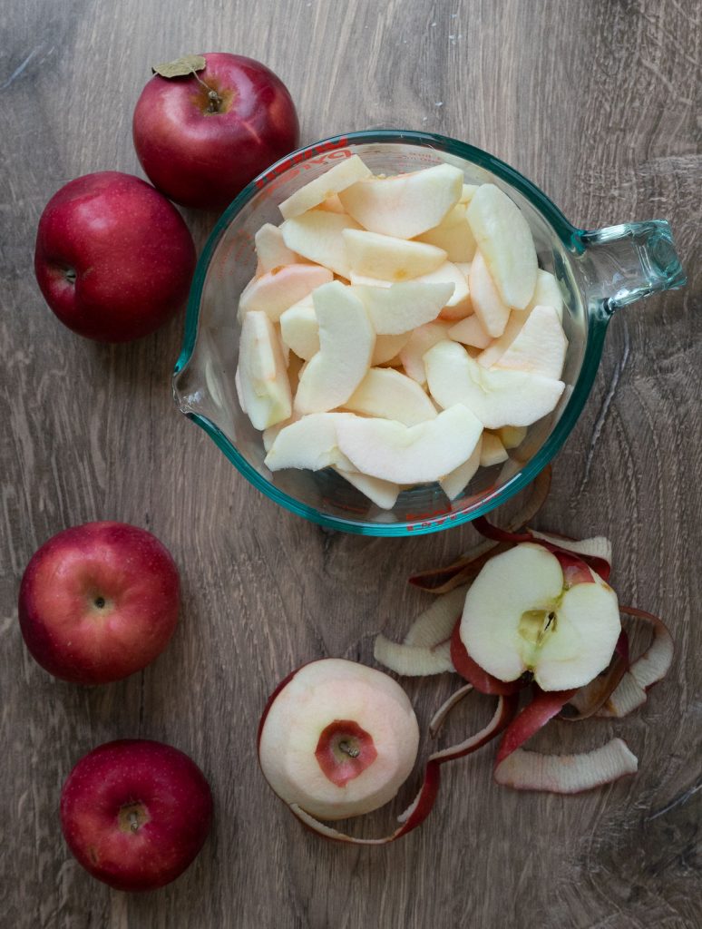 sliced apples in a bowl with apples and apple peels surrounding it on a cutting board