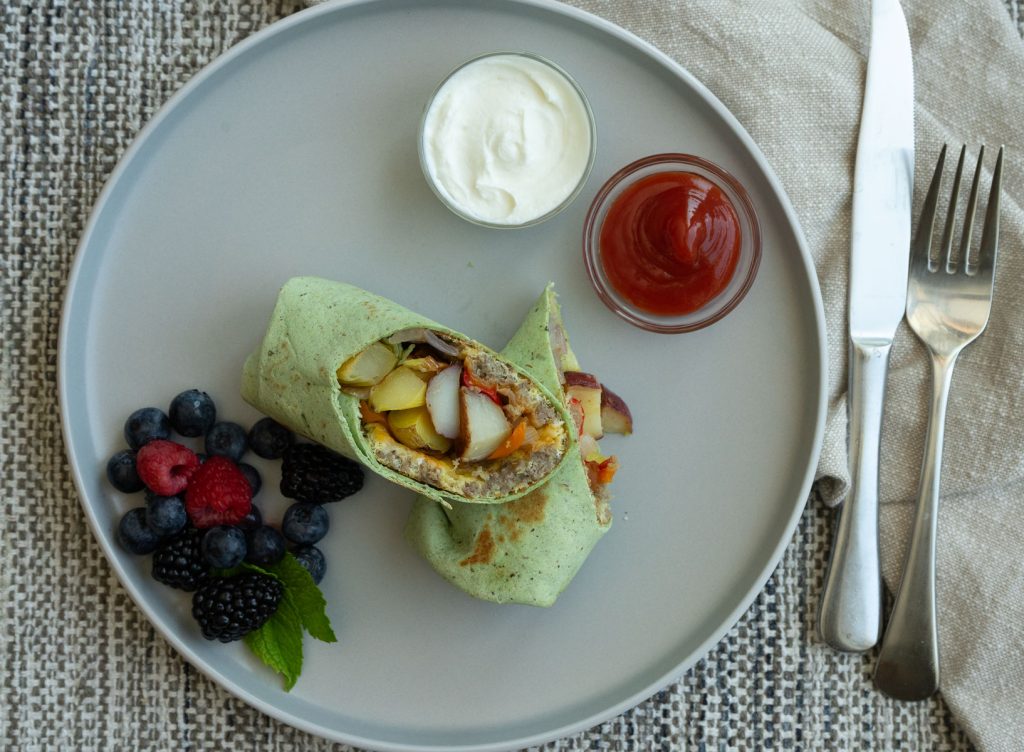 breakfast burrito in cut in half on a gray plate with berries and sour cream and ketchup in small glass cups tan napkin is to the right of the plate with a silver fork and knife