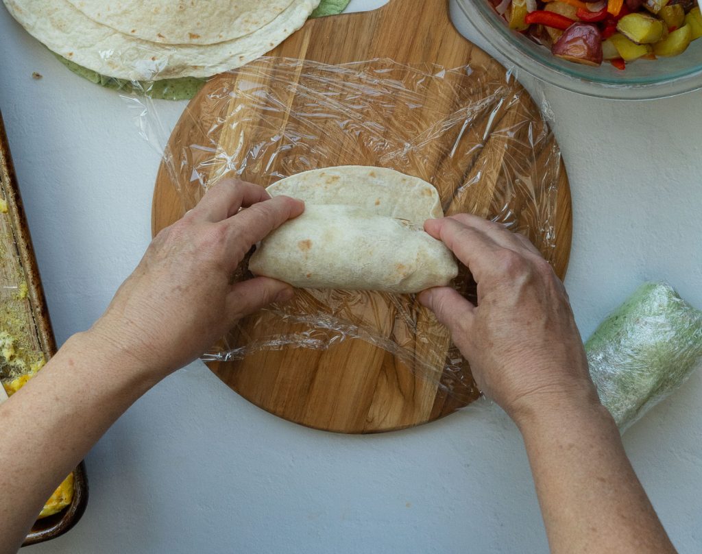 two hands rolling up a breakfast burrito, on a round wood cutting board covered with plastic wrap