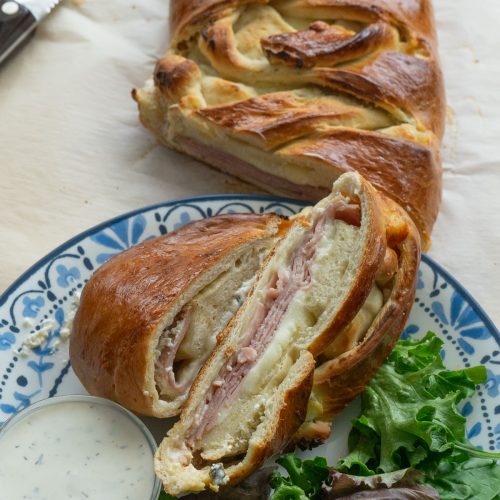 baked ham and cheese sandwich on parchment paper, slices of sandwich on a plate with ranch dressing in a small glass dish for dipping